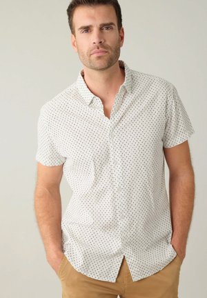 Chemise à manches courtes avec boutons, de couleur blanche avec des pois noirs, dotée d'un col et d'une poche poitrine. Le tissu semble léger et texturé.