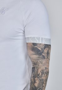 Fehér pamut póló, hímzett logóval és egy texturált, szürke csíkkal a ujján, amin a "Siksilk" felirat szerepel. Részletes tetoválás látható a karon.