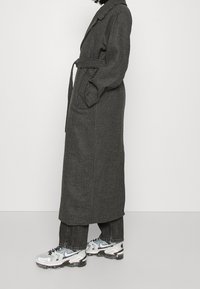 Lang zwart-wit geruite jas met een riem, zijzakken en een klassieke kraag. Gekozen boven donkere jeans en witte sneakers.