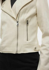 Veste en simili cuir couleur crème avec un col, fermeture éclair à l'avant et deux poches zippées. Texture lisse avec des accents en métal.
