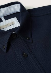 Polo bleu marine texturé avec col boutonné et boutons noirs, plié pour montrer le col intérieur rayé et l'étiquette de la marque.