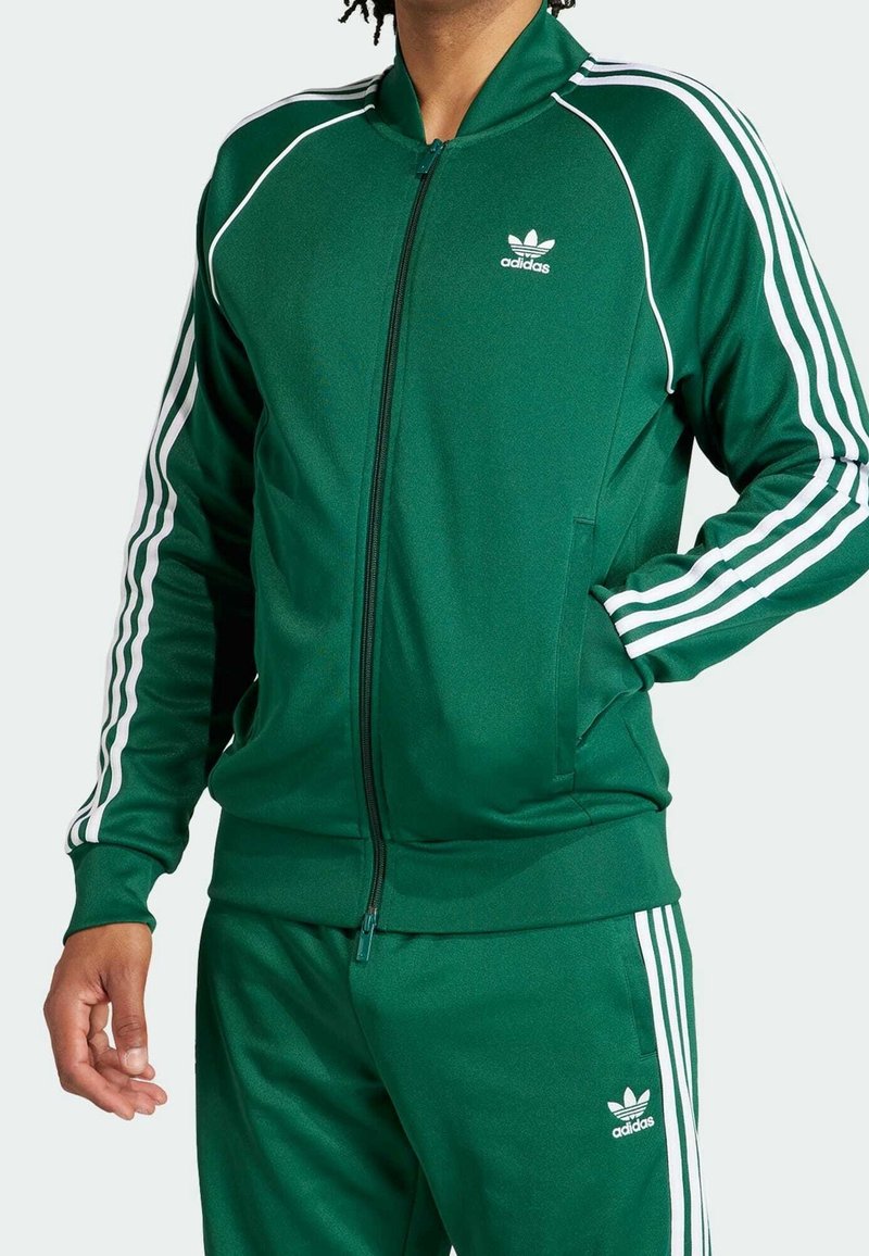 Chaqueta de chándal verde hecha de un tejido suave con rayas blancas a lo largo de las mangas y un pequeño logo blanco en el pecho. Cierre de cremallera y bolsillos laterales.