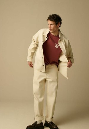 Junger Mann, der eine aufgeknöpfte beige Jacke über einem kastanienbraunen T-Shirt mit Grafikmuster richtet, passende beige Hosen und schwarze Schuhe trägt und vor einem schlichten Hintergrund steht.