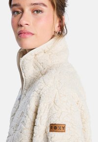 Beesidist fleece-pullover ebaõrnus tooni, millel on tekstureeritud pind, kõrgem kaelus ja tan värvi nahkpatch "ROXY" logoga varrukal.