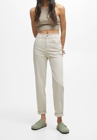 Femme portant un pantalon beige taille haute retroussé aux chevilles, des chaussures à enfiler vert olive et un crop top sans manches taupe sur fond blanc.
