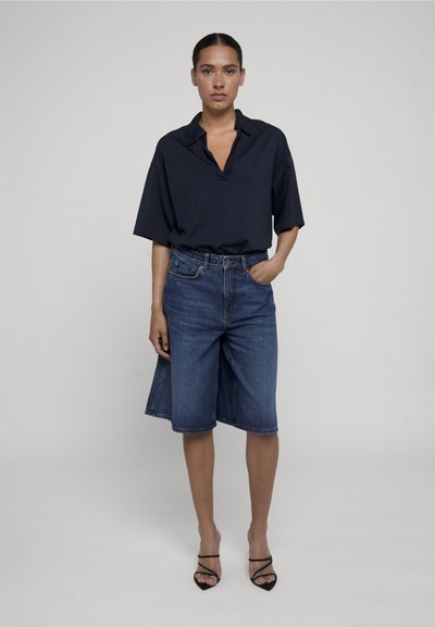 Femme aux cheveux foncés attachés en chignon, portant une chemise ample bleu marine foncé, un short en jean longueur genou et des talons noirs ouverts à l’avant, debout devant un fond uni.