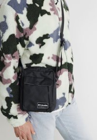 Borsetta a tracolla nera con tasca frontale e patta, realizzata in tessuto resistente, indossata sopra un giacca in pile multicolore con un motivo camouflage.