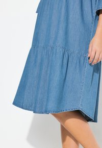 Vaaleansininen farkkumekko, jossa on kerroksellinen röyhelöhelma, lyhyet hihat ja rento malli, joka esittelee pehmeän, sileän tekstuurin.