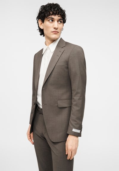 Καφέ κοστούμι προσαρμοσμένο, κατασκευασμένο από υφασμάτινη υφή, με σακάκι μονόκουμπο, γιακάδες με εγκοπή και παντελόνι στενής εφαρμογής, σε συνδυασμό με λευκό πουκάμισο.