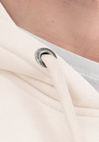 Crèmekleurige capuchontrui met een zilveren ring, voorzien van een trekkoord van dik geweven materiaal. Close-up van de stoftextuur en ontwerpdetails.