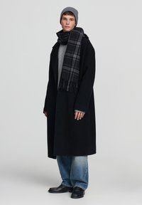 Czarny wełniany płaszcz, szara dziana czapka, szary sweter, szal w kratę, niebieskie dżinsy o szerokich nogawkach, czarne skórzane buty; model stojący na jednolitym tle.