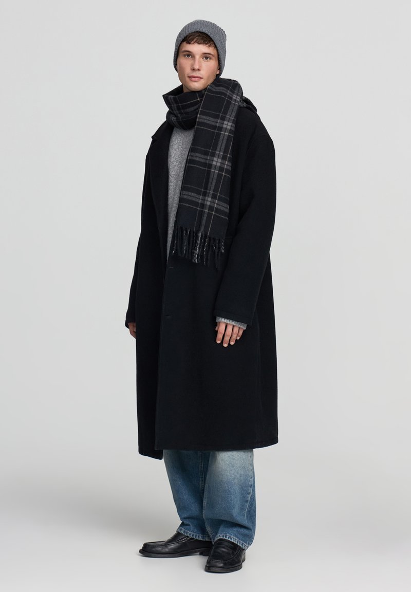 Czarny wełniany płaszcz, szara dziana czapka, szary sweter, szal w kratę, niebieskie dżinsy o szerokich nogawkach, czarne skórzane buty; model stojący na jednolitym tle.