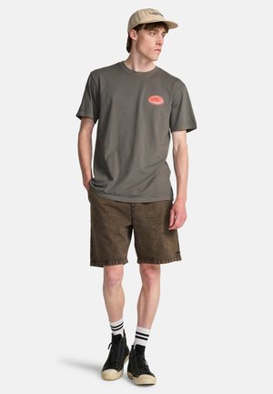Giovane uomo con una t-shirt grafica grigia, pantaloncini marroni, sneakers nere alte, calzini a righe e un cappellino da baseball beige che guarda di lato.