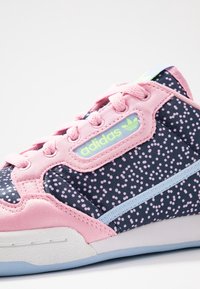 Tenisová bota Adidas s růžovým tělem, tmavě modrými a růžovými puntíkovanými detaily, strukturovanou tkaninou a světle modrým pruhem. Zelené logo je také viditelné.