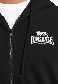 Zwarte rits hoodie van een katoenmix. Voorzien van een witte leeuwlogo en het merk "Lonsdale London" op de borst. Getextureerde voorrits.