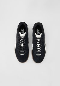 Fekete velúr sportcipő fehér díszítéssel, alacsony fazon ú, fűzős záródással és gumitalppal. A márkajelzés látható a talpbetéten.