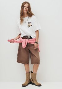T-shirt blanc avec un graphisme "animal de compagnie numérique", short en jean marron, pull rose noué à la taille et bottines beiges à chevilles avec des lanières.