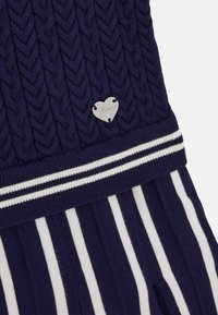 Marinblå stickad tröja med ett kabelmönster, försedd med en silvrig hjärtlogga och ett randigt vitt band vid nederkanten. Strukturdesign med ribbad finish.