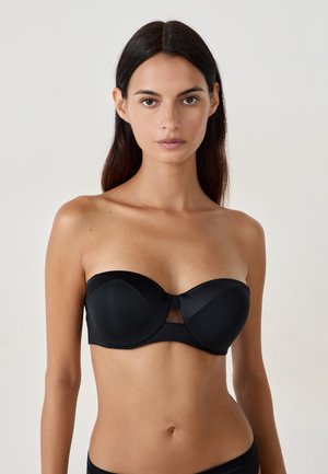 Donna con capelli lunghi e scuri che indossa un reggiseno nero senza spalline e slip neri su uno sfondo chiaro e uniforme.