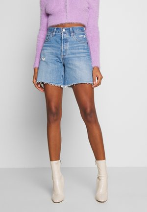 Person iført en lilla cropped sweater, højtsiddende distressed denimshorts og hvide ankelstøvler, der står mod en ensfarvet baggrund.