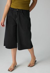 Pantalones culotte de mezclilla negra con un ajuste holgado, cintura de media altura y diseño de cinco bolsillos. Combinados con una blusa verde texturizada y sandalias con estampado.