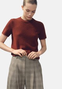 Pull en maille à manches courtes couleur rouille, associé à un pantalon taille haute à carreaux gris clair, avec une coupe décontractée et une texture de tissu visible.