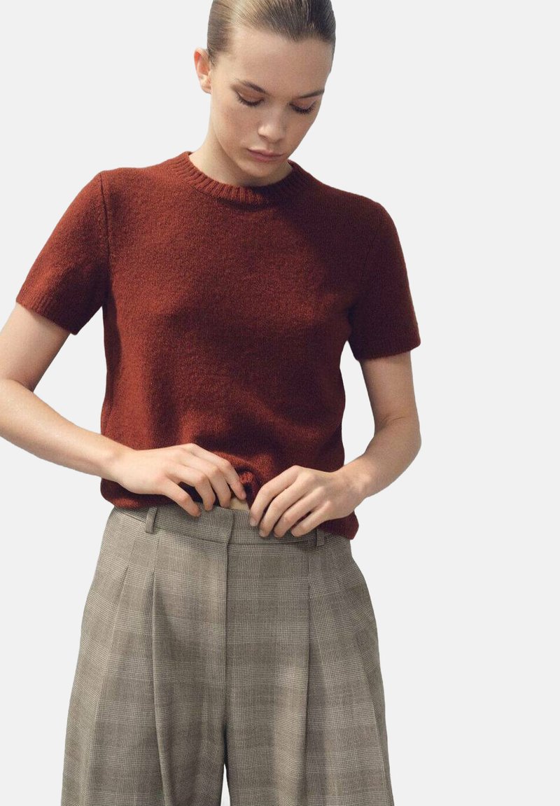 Pull en maille à manches courtes couleur rouille, associé à un pantalon taille haute à carreaux gris clair, avec une coupe décontractée et une texture de tissu visible.