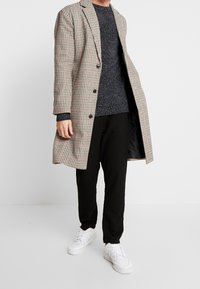 Kostkovaný dlouhý kabát v béžové, černé a červené barvě, zkombinovaný s tmavým strukturovaným svetrem a černými kalhotami, doplněný bílými teniskami.