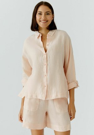 Lächelnde Frau, die ein hellrosa Hemd mit Knöpfen und langen Ärmeln sowie passende Shorts trägt, vor einem einfarbigen hellen Hintergrund.