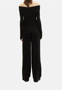 Femme portant un haut noir à épaules dénudées à manches longues et un pantalon noir à jambes larges, debout de dos sur un fond uni.