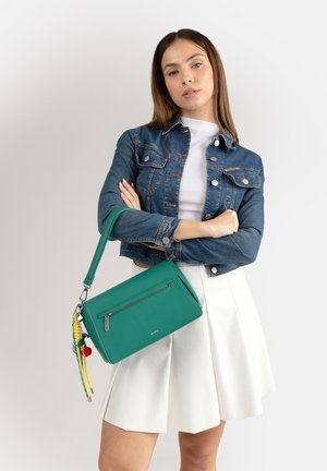 Donna con giacca di jeans e gonna bianca che tiene una borsa verde con nappine colorate su sfondo semplice.