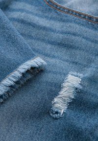 Ljust blått denimtyg med fransad fåll, slitna lappar med lösa trådar och synlig sömnad i en kontrasterande orange färg.