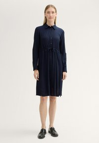 Abito navy con bottoni, vita con cinturino, maniche lunghe e orlo a ginocchio. Tessuto morbido con texture sottile e spacchi laterali per facilitare il movimento.