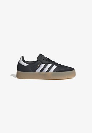 Černá teniska Adidas Samba s bílými pruhy, gumovou podrážkou a zlatým nápisem „SAMBA“ na boku, zobrazena z boku na bílém pozadí.