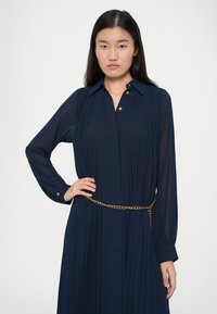 Γυναίκα ντυμένη με ένα ναυτικό μπλε πλισέ φόρεμα με χρυσά κουμπιά και μια χρυσή ζώνη, ποζάροντας με το ένα χέρι στη μέση της σε λευκό φόντο.