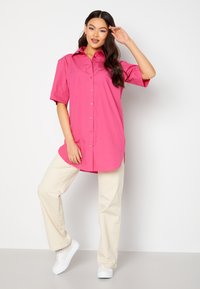 Camicia corta rosa brillante con design a bottoni, abbinata a pantaloni a gamba larga beige chiaro e sneakers bianche. Texture liscia.