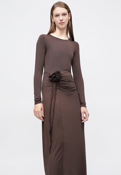 Γυναίκα που φοράει μακρυμάνικο, καφέ, φόρεμα μέχρι το πάτωμα με σούρα στη μέση και διακοσμητικό υφασμάτινο λουλούδι, στέκεται μπροστά σε μονόχρωμο φόντο.