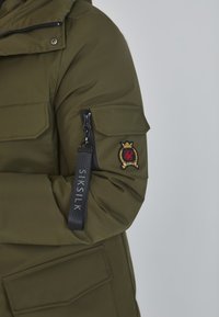 Olivovo zelená parka s vreckom na zips, nášivkou na pleci a značkovou etiketou. Obsahuje kapucňu a nastaviteľné manžety. Hladká, textúrovaná látka.