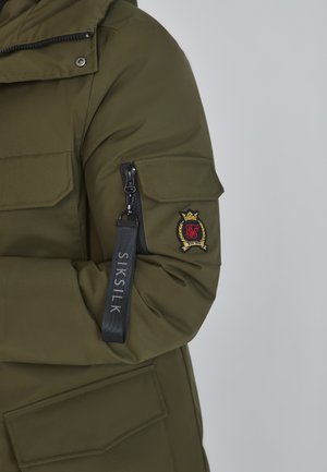 Parka de color verde oliva con un bolsillo con cremallera en la manga, logo bordado en el brazo y una correa que dice "SIKSILK."