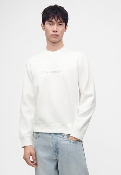 Νέος άνδρας που φοράει μια λευκή φούτερ Emporio Armani και ανοιχτόχρωμο μπλε τζιν, στέκεται μπροστά σε ένα απλό λευκό φόντο.