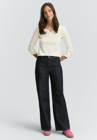 Donna con capelli castani mossi indossa una maglietta henley crema a maniche lunghe, jeans larghi neri e sneakers rosa, in piedi con le mani dietro la schiena.