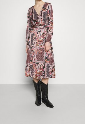 Kobieta ubrana w sukienkę midi z wielokolorowym wzorem paisley i geometrycznym, połączoną z czarnymi kowbojskimi kozakami za kostkę, stojąca na szarej podłodze.