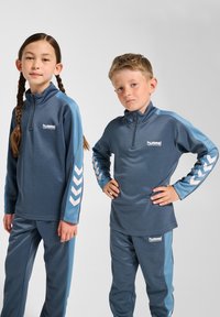 Twee kinderen dragen bijpassende sportoutfits van glad, donkerblauw stof met lichtblauwe accenten en chevronpatronen op de mouwen.