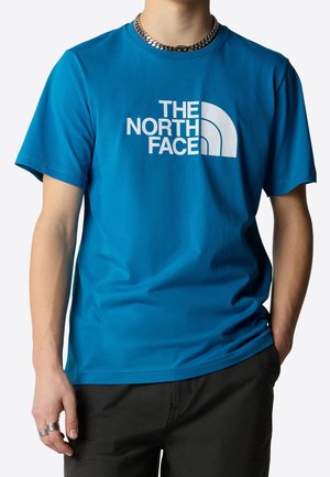 Mężczyzna w niebieskim t-shircie z krótkim rękawem z białym logo "The North Face" i czarnych spodniach, jedną ręką w kieszeni, srebrny łańcuszek i pierścionki.