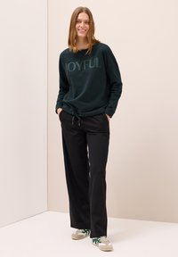 Mörkgrön sweatshirt med texten "JOYFUL", i kombination med svarta löst sittande byxor. Modellen bär beige och gröna sneakers. Enkel bakgrund.