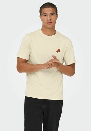 Cream-colored short-sleeve T-shirt featuring a small red and yellow graphic on the chest. Paired with dark jeans. Simple design and fit.