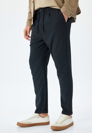 Mørkeblå stribede bukser med snoreliv, sidelommer og tapered ben, parret med cremefarvede sneakers og brune gummisåler.