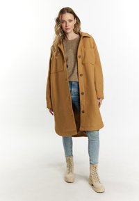 Lungo cappotto color cammello con chiusura a bottone e due tasche sul petto, abbinato a un maglione beige e jeans azzurri chiari, con stivali beige con lacci.