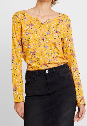 Kobieta w żółtej, długorękawowej bluzce w kwiaty, włożonej do wysokiej talii czarnej spódnicy z denimu z guzikami i kieszeniami.