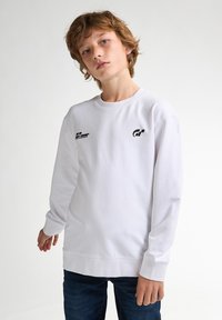 Sudadera blanca de manga larga en algodón con cuello redondo. Presenta un logotipo negro en el pecho y un corte relajado. Combinada con jeans azules.
